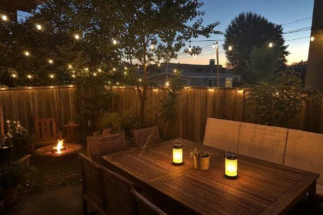 A small fenced backyard at dusk with string lights overhead, a lit fire pit and two glowing lanterns on a wooden table.