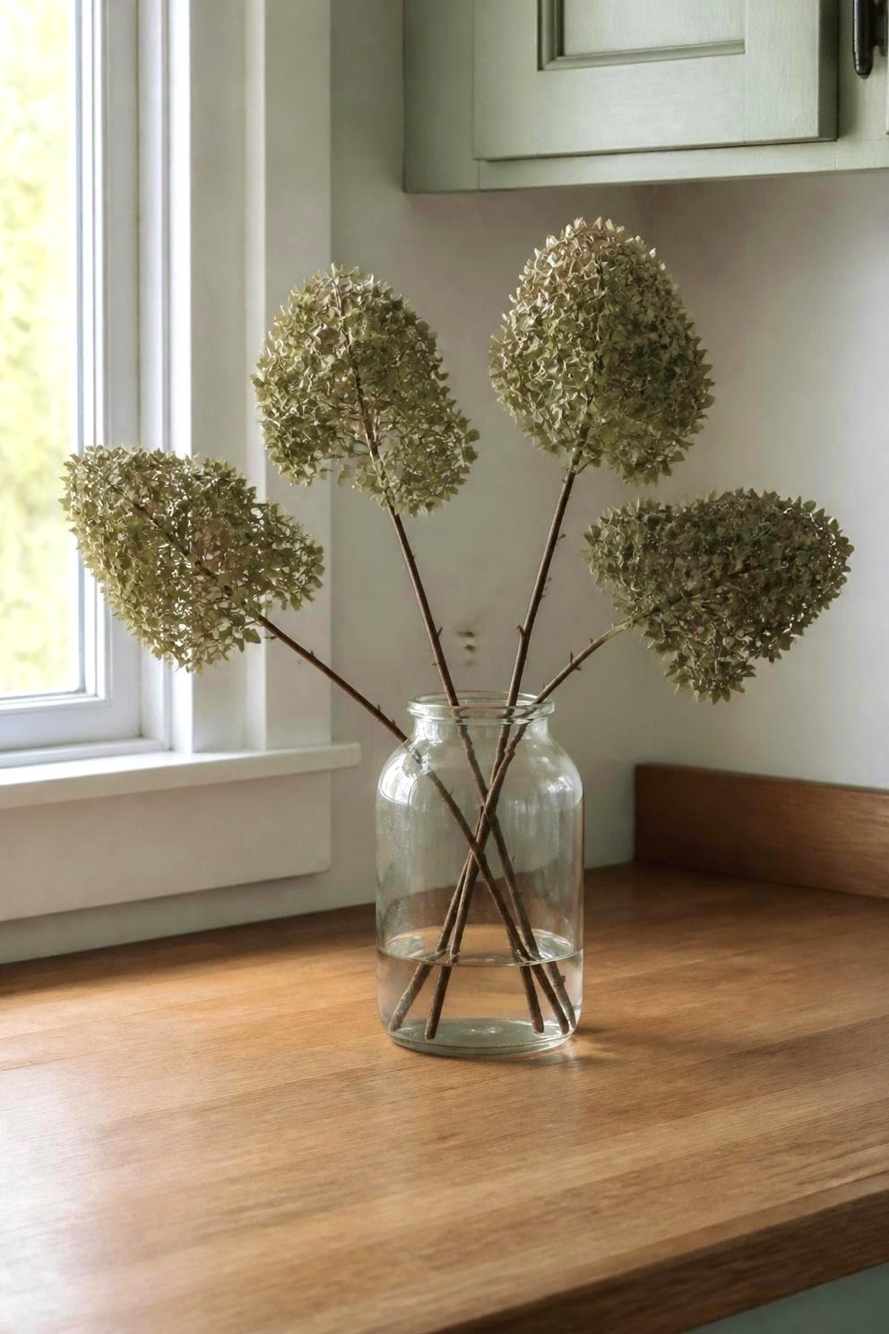 Four dried panicle hydrangea stems of different heights in a clear glass jar on a kitchen counter by a window