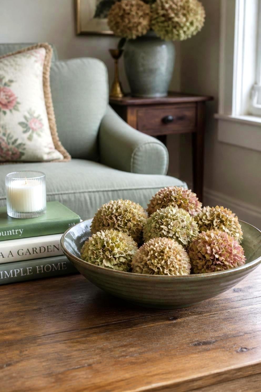 A shallow green bowl of dried hydrangea heads on a coffee table beside a candle and books