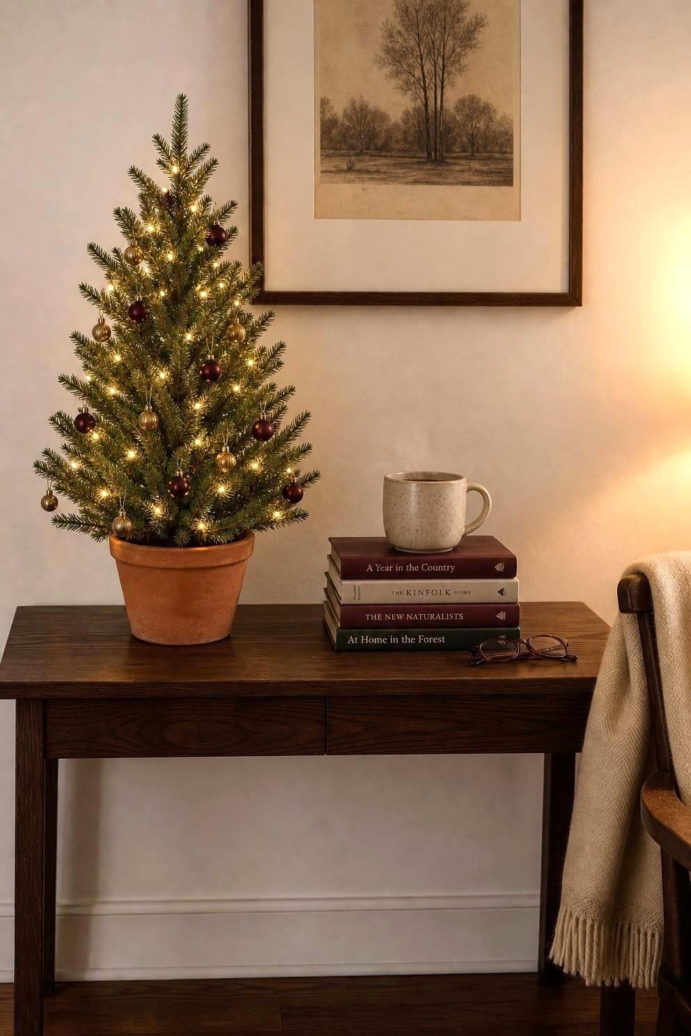 Small lit Christmas tree in a terracotta pot on a dark wood console beside a stack of books and a mug