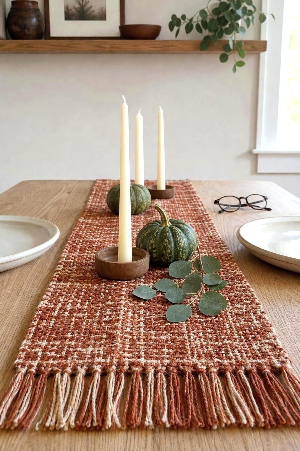 A narrow rust and cream table runner laid down the center of a small dining table with three unlit ivory taper candles in low wooden holders and two small green gourds beside them