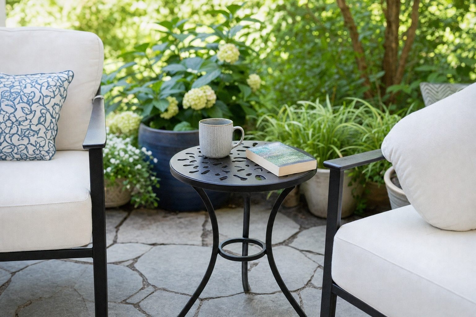 Small round dark metal patio side table with a cut-out top standing between two outdoor chairs, holding a mug and a book