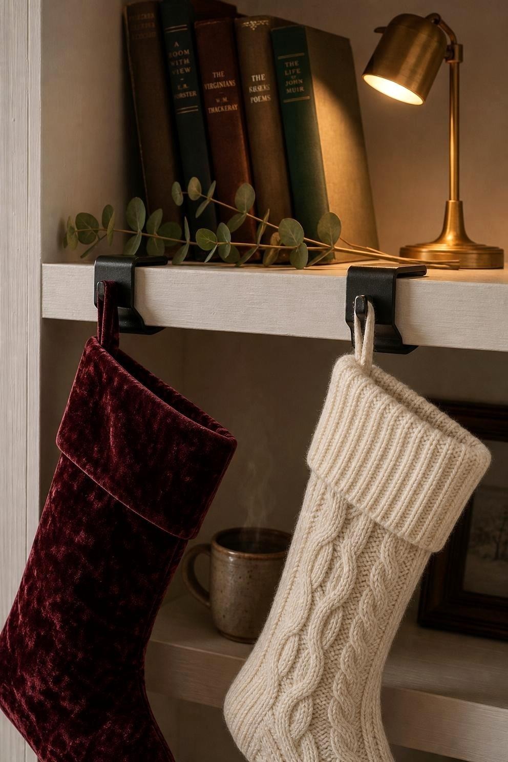 Two knitted stockings hanging from black clamp holders gripping the edge of a wooden bookshelf, with old books, eucalyptus and a small brass lamp above